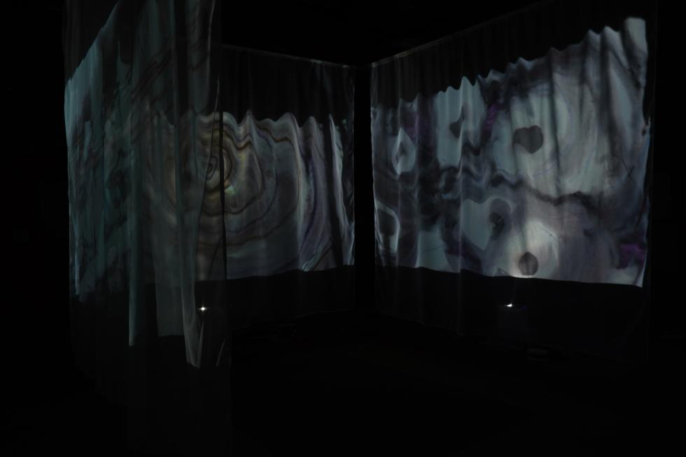 Abstract images resembling ripples in water are projected onto floating fabric screens in a dark, black room. 