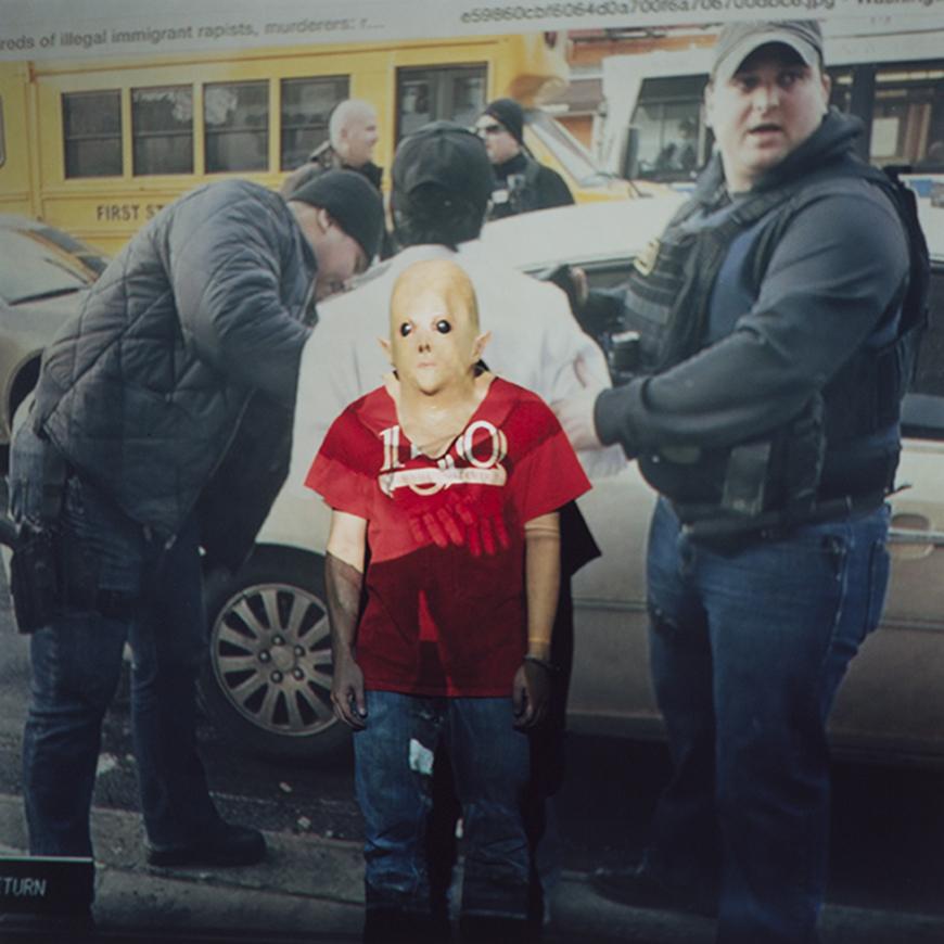 Color photo of a person in a red tshirt and blue jeans, wearing a plastic masks of a bald, light skinned face, with a projection over the subject of the photo. The project is two police officers escorting a handcuffed person into a car, with school buses and additional police in the background. 