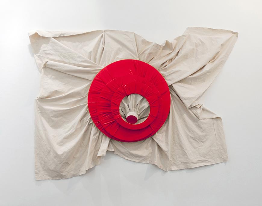 Red colored circular fabric with a hole cut in the middle on top of a white sheet pinned to the wall.