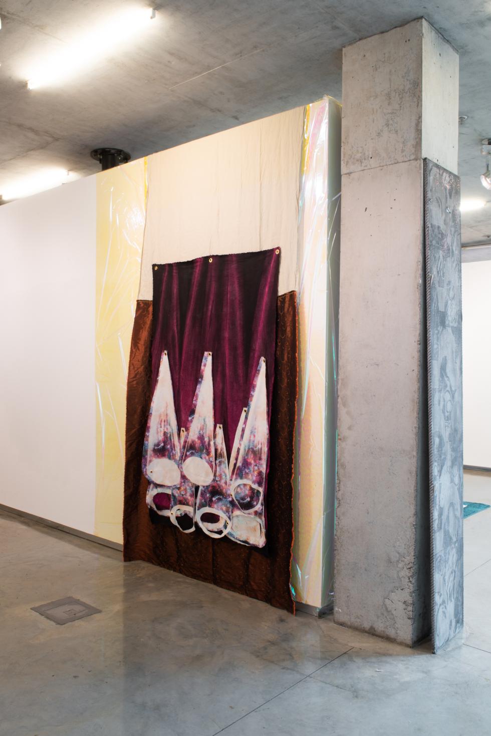 A large piece of maroon-colored cloth hangs on a white gallery wall next to a concrete pillar, with plastic and vinyl between the cloth and the wall. Painted on the fabric are spotlight motifs in a mix of white, purple, and pink. 