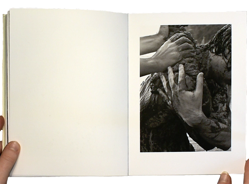 Scanned photo of a handmade photo book. Left page is blank; on the right page is a black and white photograph of a person embracing themselves while being covered in mud by another's hands. The photo is printed on cream colored pages with thick margins.