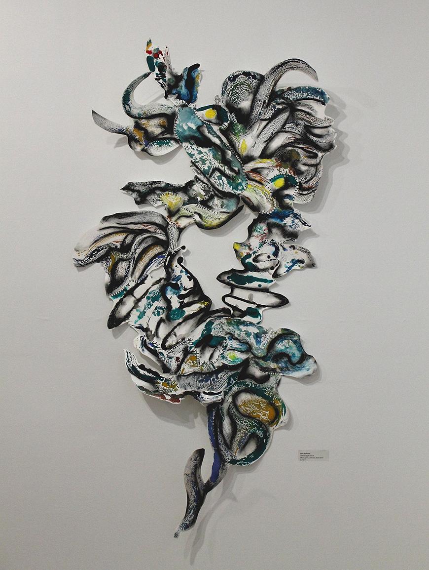 Color photo of an abstract mushroom-like sculpture made of black, green, blue, and yellow watercolor on white paper, hung on a white wall.