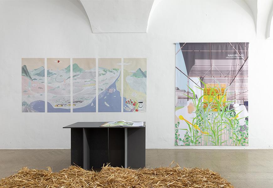 An art gallery display featuring two large artworks on a white wall. On the left, there are five panels depicting a scenic landscape. On the right, there is a single piece showcasing a modern structure with green plants in the foreground. A dark, minimalist table stands in front of the artworks, with a book or catalog open on it. The floor around the table is covered with straw.