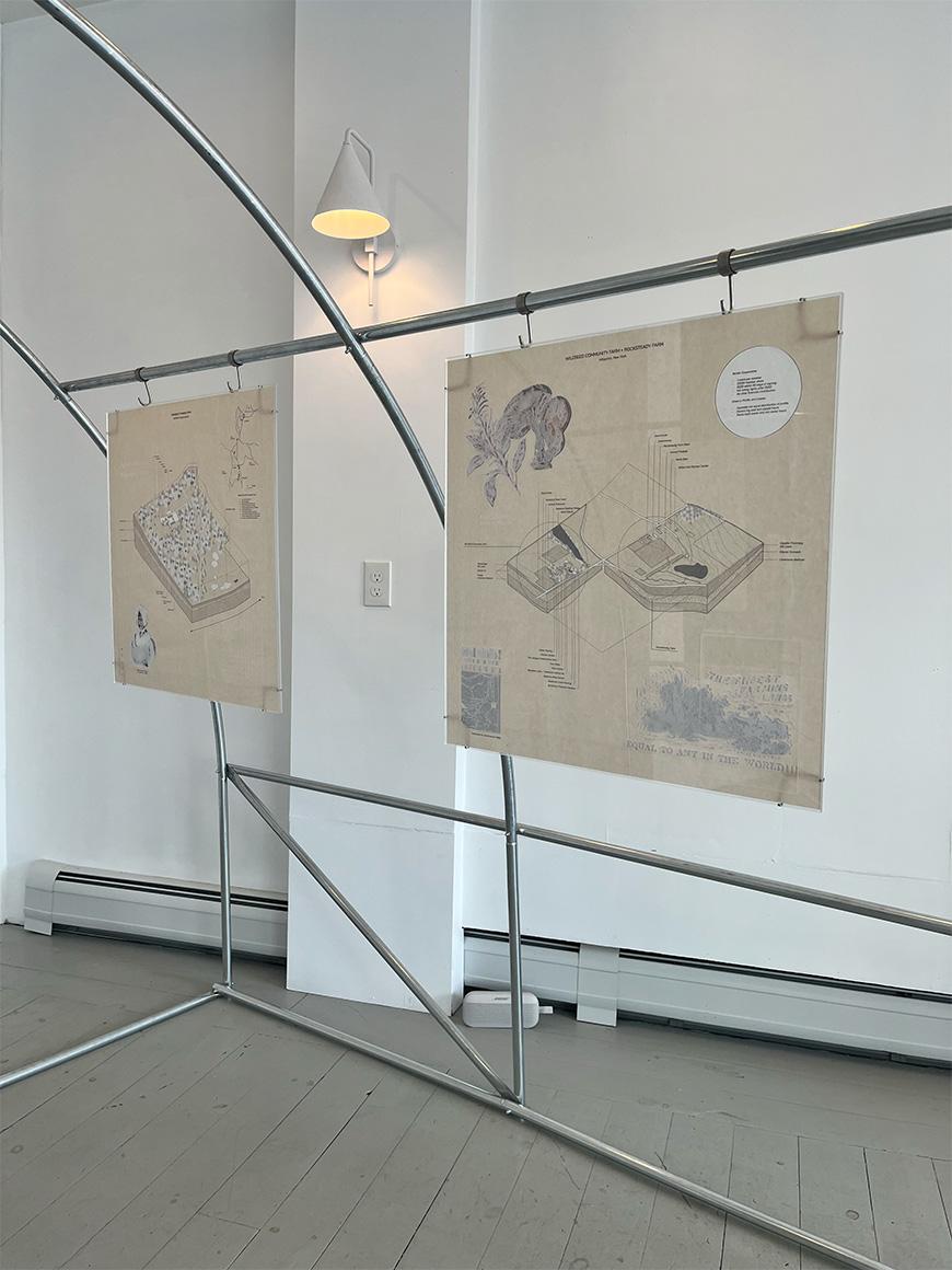 An art installation in a gallery featuring two detailed drawings on beige paper, hung from a metal frame structure. The drawings depict intricate diagrams and illustrations. A minimalist wall lamp is illuminated in the background, casting a soft light over the exhibit. The gallery space is bright and clean with light-colored wooden floors.