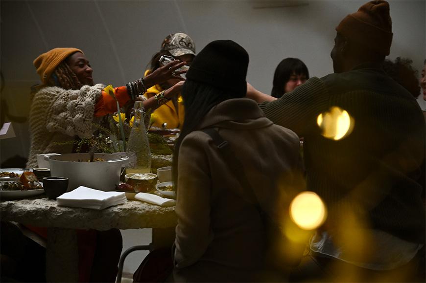 A group of people gathered around a hempcrete table enjoying themselves and passing around a bottle. The atmosphere is dimly lit and cosy, and the table is filled with food and floral arrangements.