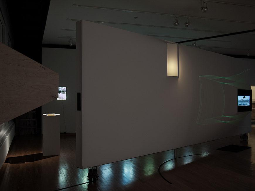  A dimly lit gallery space with a large, white wall dominating the center. The wall has a small, rectangular light fixture near the top. On the left side of the wall, a large, angular sculpture casts a shadow. In the background, there is a video screen displaying a nature scene.