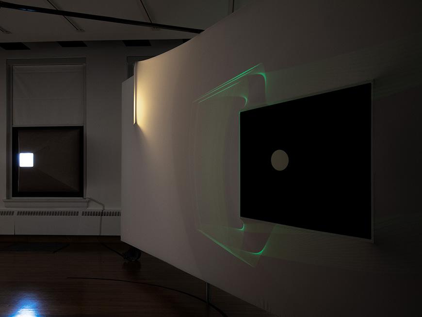 A dark, minimalist art installation featuring a large white curved wall with a black square in the center. The square has a glowing green circle inside of it, and there are faint green lines projecting from the circle onto the wall. The room is dimly lit, with a single light source casting a shadow on the wall.