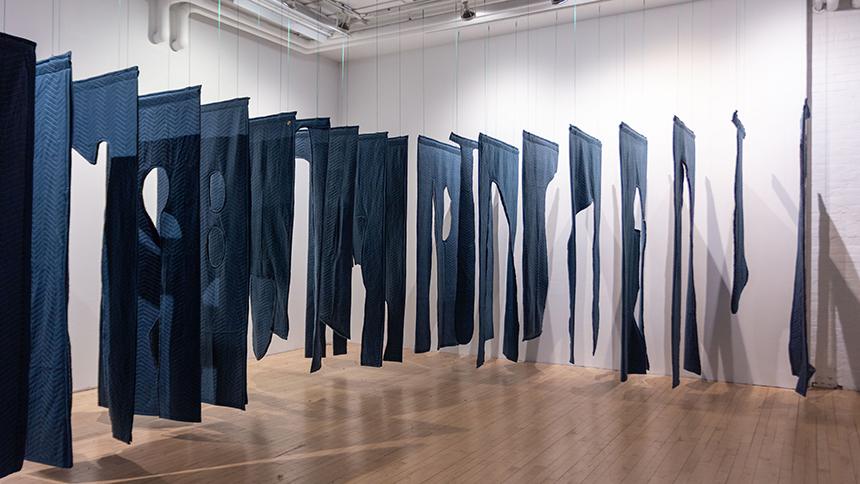 Panels of blue fabric cut in various shapes hang from the ceiling, nearly touching the floor. The room is brightly lit with white paneled walls and a pale wood floor.