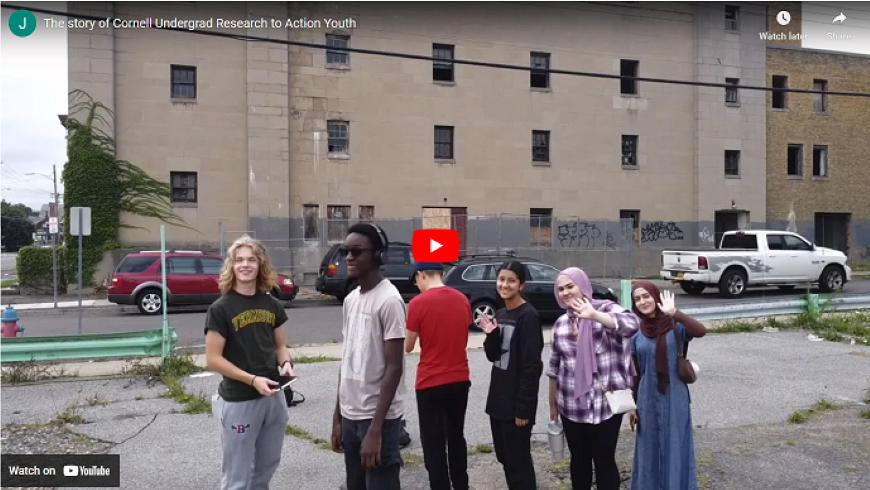 A group of six students waving at the camera with a graffitied building in the background