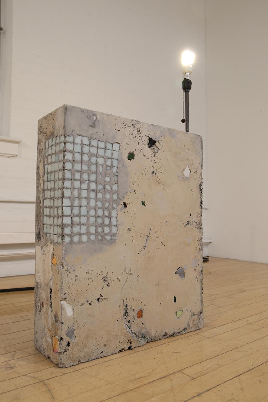 Cinder block with a lit light bulb on top with random objects cemented together.