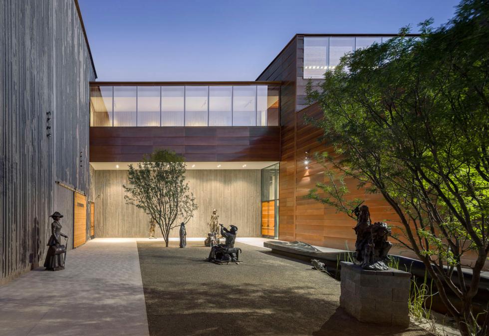 A modern wood and glass building lit up at dusk. there is a concrete and gravel courtyard with sparse foliage and statues about