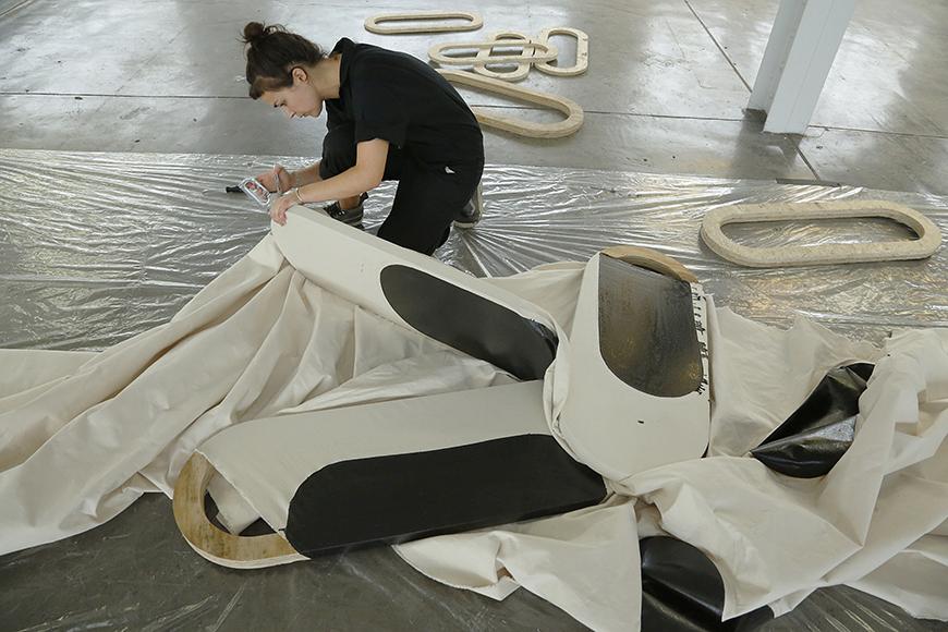 Artist stapling white cloth with black shapes painted on it to a wooden oblong shape.
