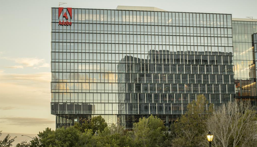 Large office building with Adobe sign on outside of building. The building is covered with mirrored glass and there are trees in the backround. 
