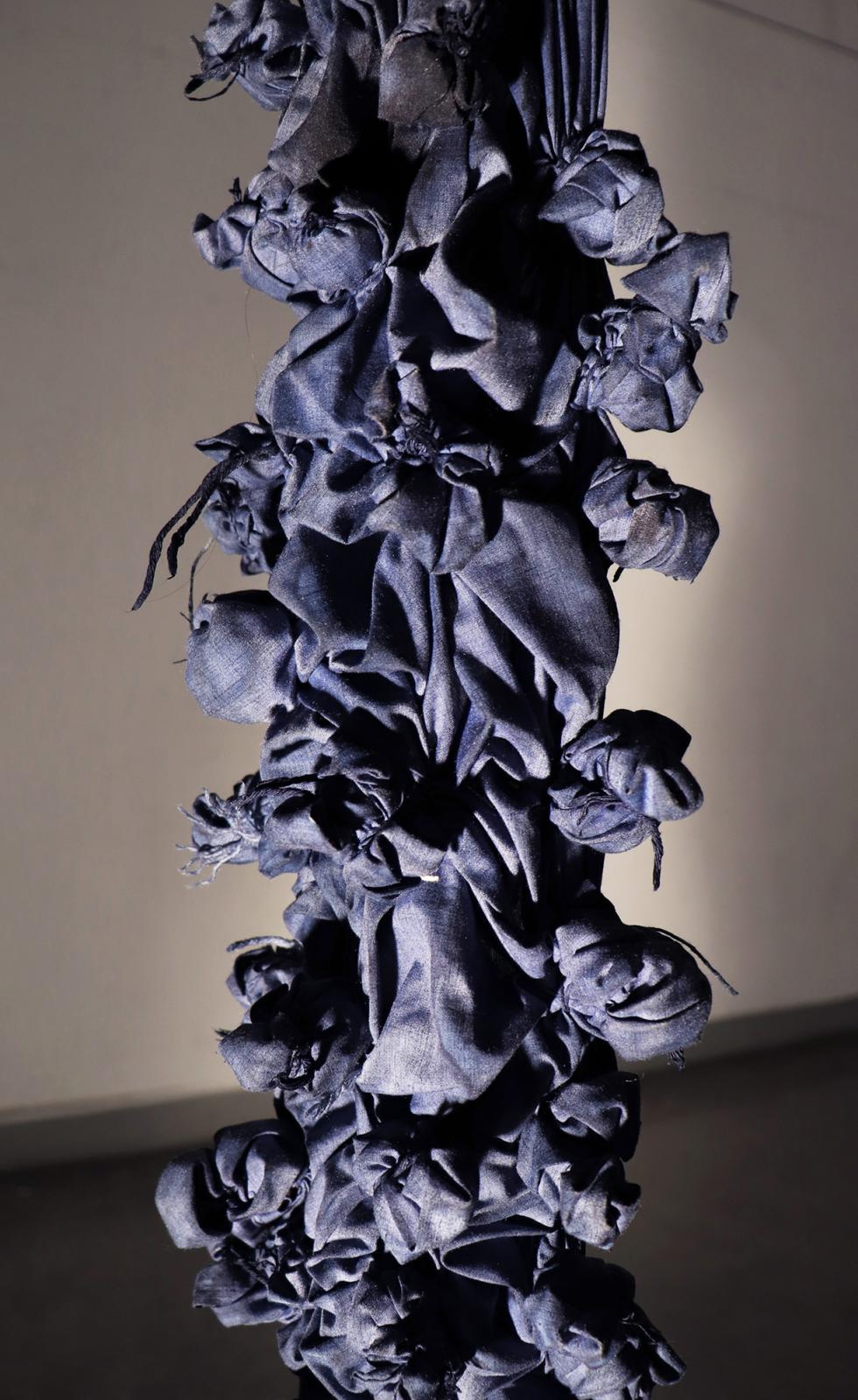 Close up of pieces of dark purple cloth bound together with dark purple yarn in bunched up patterns hung from the ceiling.