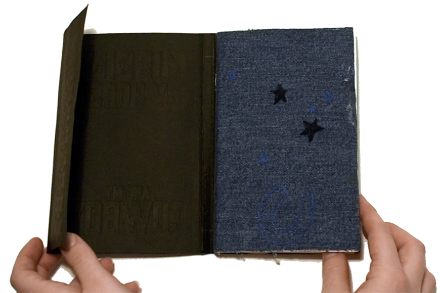 Light skinned hands hold open a book so that the covers are visible. The cover is made from dark blue denim and brown material which is reminiscent of suede. Black star shapes are on the blue denim side. 