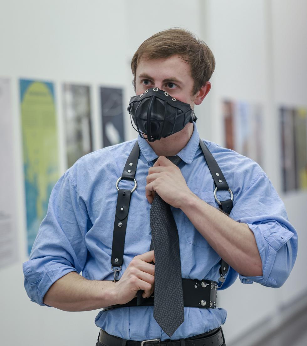 Blonde white man dressed in a blue-collar t-shirt wearing a black leather harness and with metal rings and a leather-looking maskwith a speaker in place of its mouth.