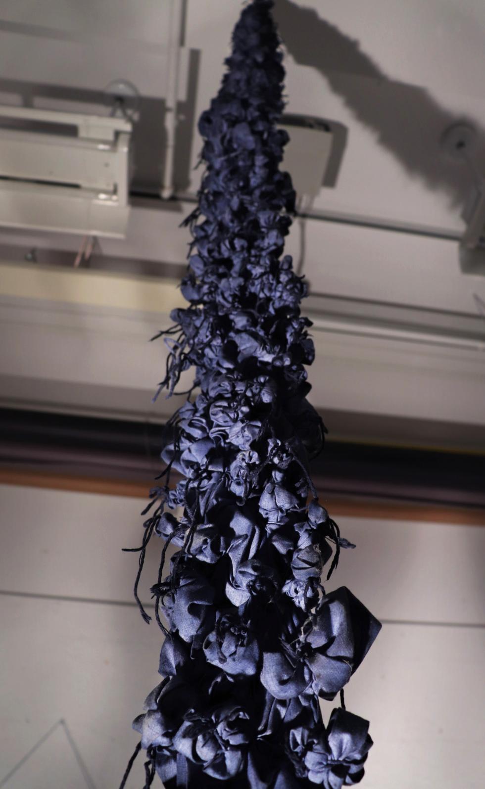 Pieces of dark purple cloth bound together with dark purple yarn in bunched up patterns hung from the ceiling.