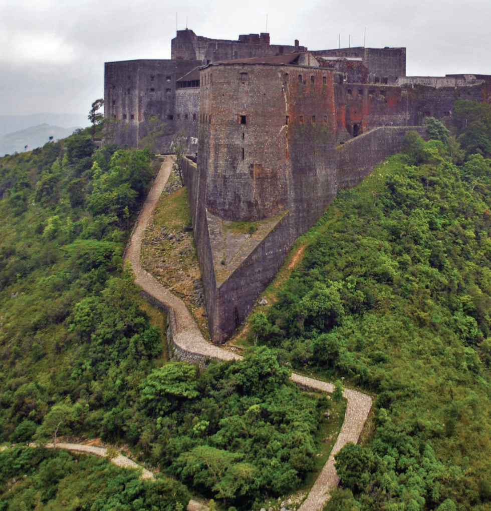 A massive, stone citadel perched on a hilltop overlooking lush greenery. A winding path leads up to the fortress, which features thick walls, towers, and a large central structure.