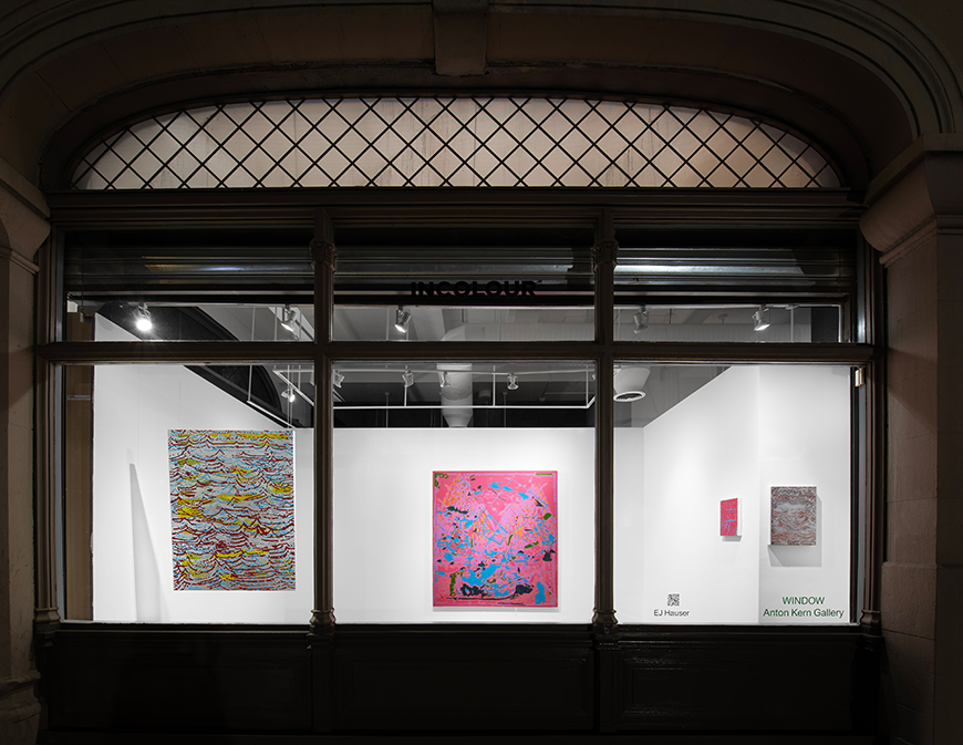 Multicolored abstract paintings in a white-walled gallery, on view through a glass window. 