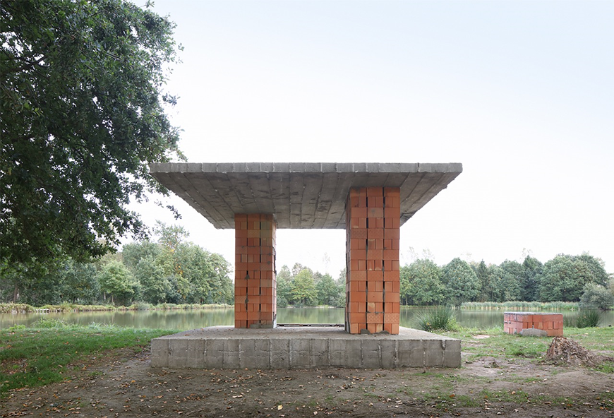 A small, minimal pavilion constructed from cement and two brick pillars. The pavilion is situated next to a pond. 