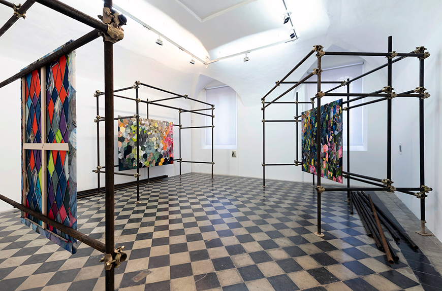In a room with white walls and a checkerboard floor sits three sculptures made out out metal iron pipe scaffolding which multicolored quilts hang from. 