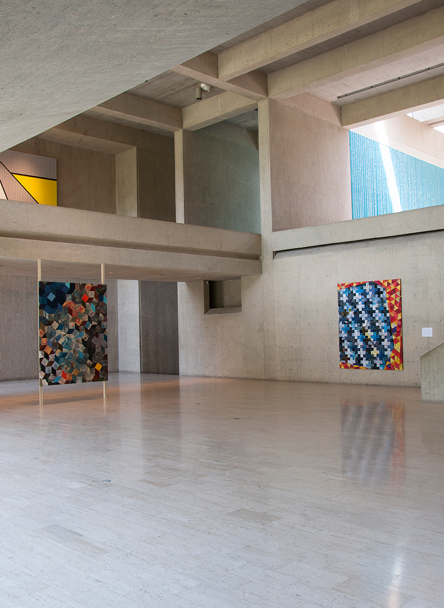 In a gallery with light grey concrete geometric architecture hangs two brightly colored multi-colored quilts in an otherwise empty space.