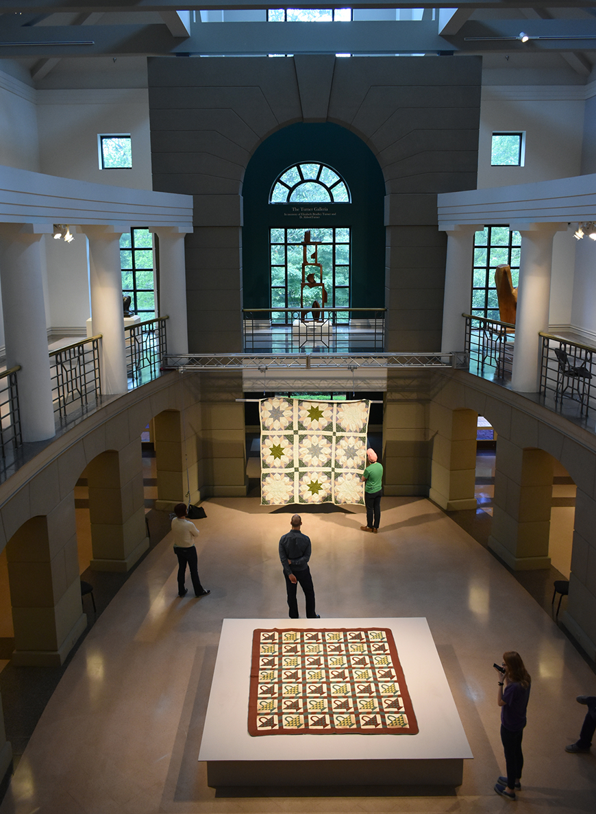 Multiple people stand in a two-story room with multiple quilts on display in it, with a balcony overlooking the first floor's quilts and sunlight coming in through a wall of windows and a skylight. 