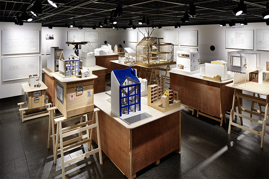 The interior of a gallery space which is occupied by a series of models at the center of the room, with the walls covered in a series of technical drawings. 