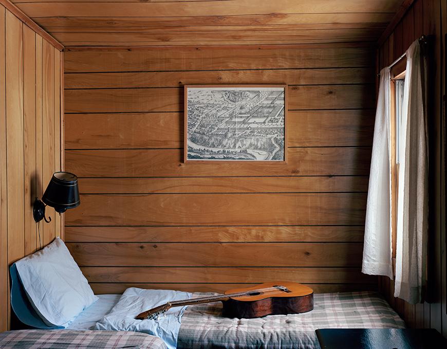 Horizontal color photo of a bed in a log cabin with a brown guitar on top of plaid blanket and baby blue bedsheets. A black sconce hangs on the wall above the head of the bed, on the left. At the foot of the bed, on the right side of the image, is a white curtain over a window letting sunlight into the frame.