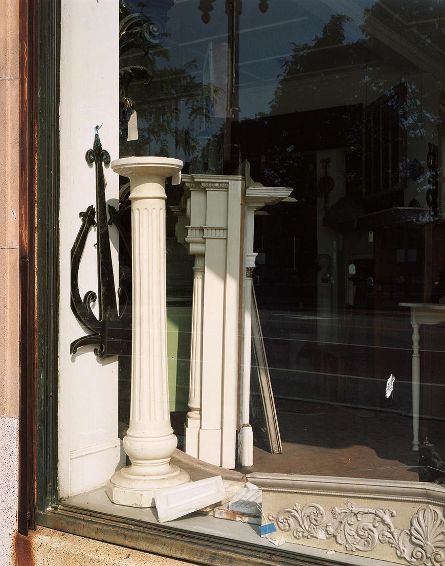 Vertical color photograph of two free standing architectural columns inside of a storefront window. The one in the background is partially shaded and the other in the foreground is lit by the full sunlight.