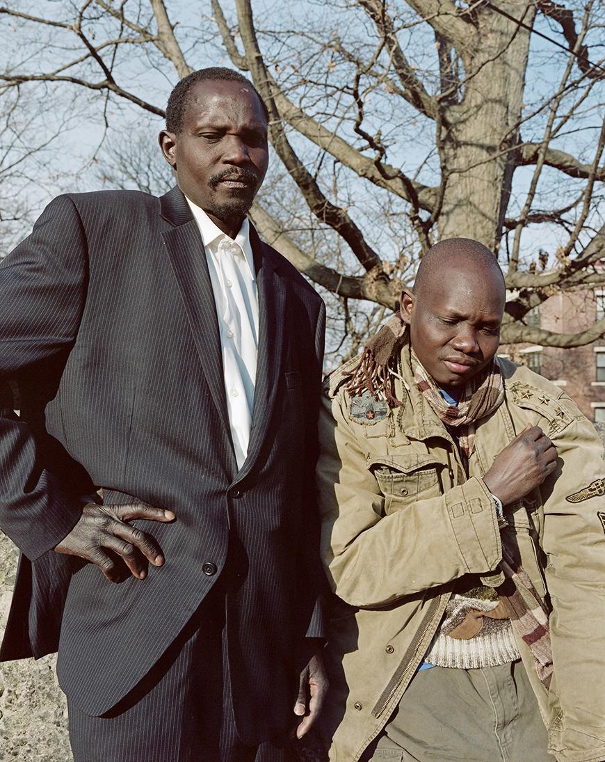 Vertical color photo of two dark-skinned individuals standing outside in the sun in front of a tree with blue sky in the background. The subject on the left has their hand on their hip, wears a black suit and white button-up, and has a black mustache and short hair. The subject on the right is bald and wears an army green coat and pants, with a striped sweater and scarf, holding their hand in a fist over their heart with eyes closed. 