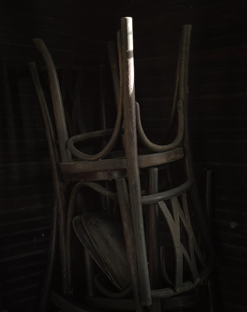 Vertical color photo of a dimly lit chair turned upside down on top of another chair. The entire photo is staged in the dark except a single leg of the upturned chair in the center of the photo. 