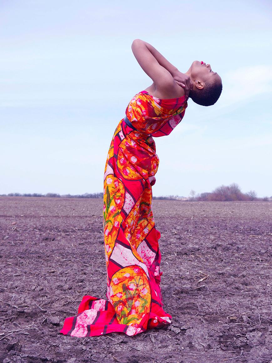 Photograph of a dark skinned person in a barren field against a blue sky background. The person stands in a bright red abstract patterned dress, leaning back with the left hand on the back of their neck, with their eyes closed and face toward the sky.