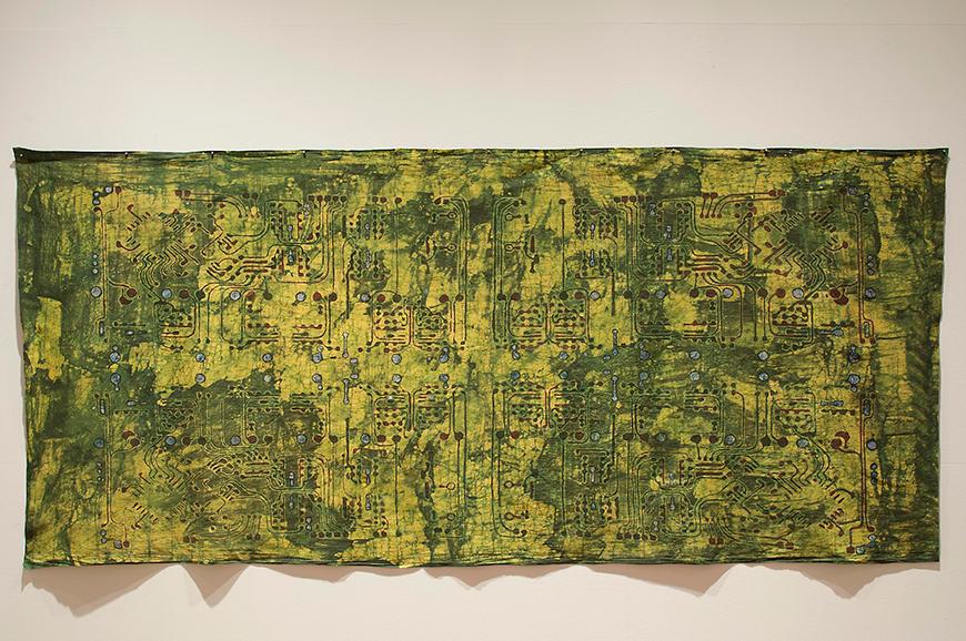 Color photograph of textile art hung on a white wall. The muslin fabric base is green and mustard colors, with black designs reminiscent of computer chips and electronic pathways painted on in black. 