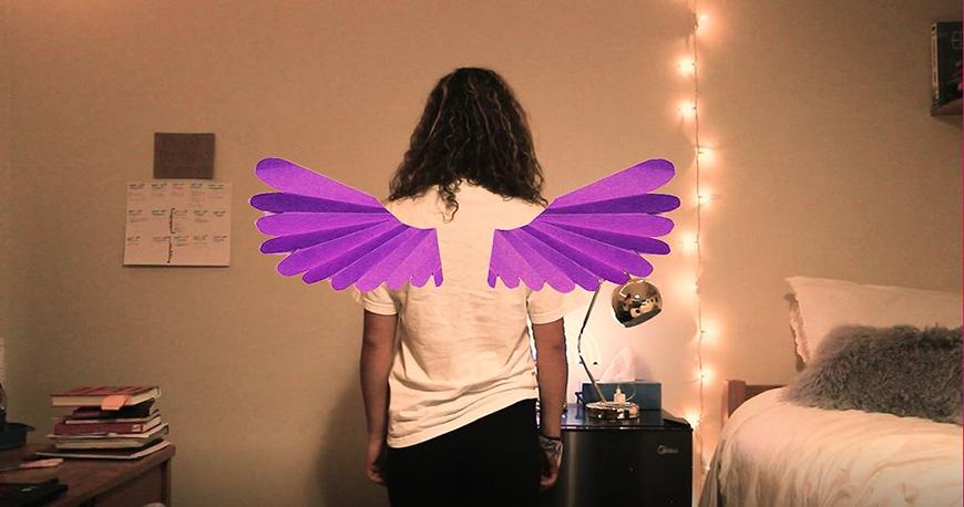 A person turned away from the camera wearing purple colored construction paper wings.