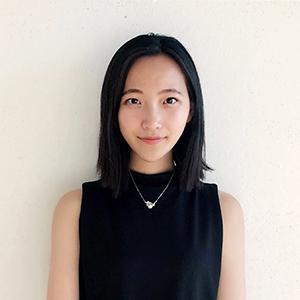 headshot of a young woman with black hair and a black sleeveless shirt standing in front of a white wall