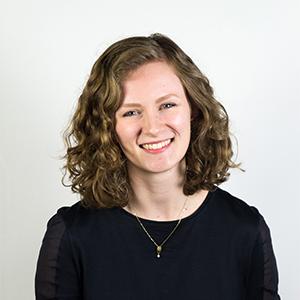 headshot of a young woman with light brown curly hair wearing a dark sweater and standing in front of a white wall