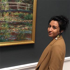 Woman wearing a tan sweater with dark hair in a bun in front of a painting in gold frame against a dark wall.