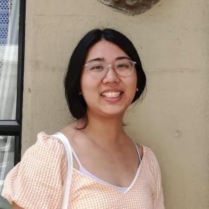 Girl with dark hair loosely pulled back wearing a light orange striped shirt against a pale tan wall.