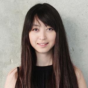 headshot of a woman with long dark hair wearing a black sleeveless shirt in front of a grey wall