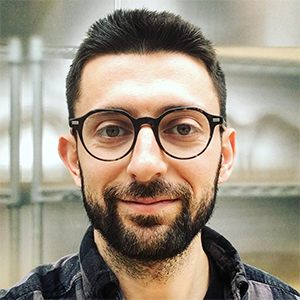 headshot of man with short dark hair and beard with shelf in background