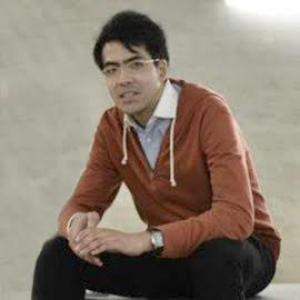 man with dark hair and glasses wearing a dark orange shirt and sitting on a bench