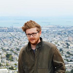 head and torso of a man with reddish hair and glasses with a cityscape behind him