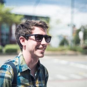 headshot of a young man in a plaid shirt wearing sunglasses with a street behind him