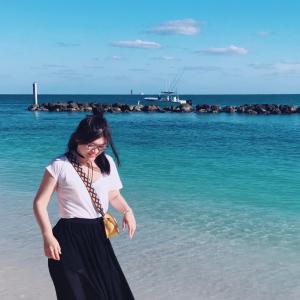 woman walking on a beach wearing a white shirt and black skirt with blue ocean and sky behind her