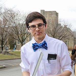 headshot of a man with short dark hair wearing a blue bow tie, glasses, and a white shirt with a building and trees behind him