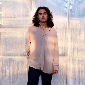 A young man with shoulder length brown hair, who is wearing a cream colored button up shirt. 