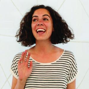 headshot of a young woman with dark curly hair wearing a black and white striped t-shirt raising one hand
