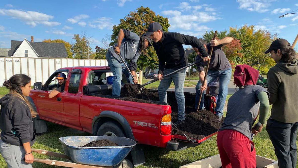 Two men in the back or a red truck bed shoveling dirt with a young woman pushing a wheelbarrow and two other students working in a raised garden ben in front.
