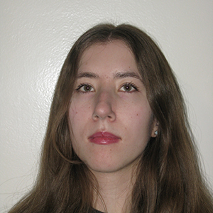 A woman with light skin and straight brown hair, standing unsmiling in front of a white background.